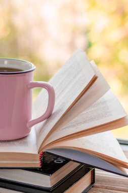 Pembe bir fincan çay açık bir pencerenin önünde açık bir kitap yığınının üzerinde duruyor.