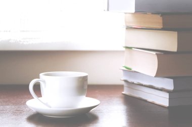 Kitapların yanındaki tahta masada beyaz bir fincan kahve duruyor.