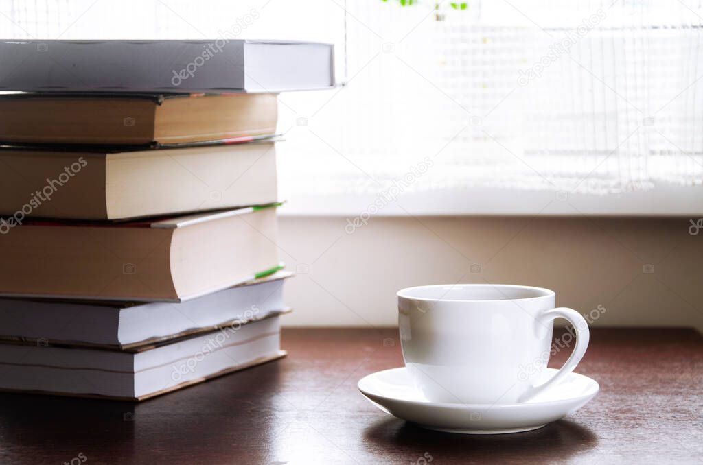 A white cup of coffee stands on a wood table next to the books