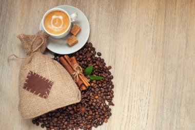 Fincan espresso kahve çekirdekleri, çuvala ile ışık ahşap zemin üzerine tarçın ile. Saat işareti ile kahve köpüğü.