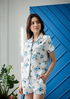 Açık mavi çiçek desenli beyaz pijama giyen, mavi duvarın önünde poz veren ve saksıdaki yeşil bitkiler için poz veren genç, zayıf, esmer bir kadın. Uyku giysisi tasarımı. Sabahları tatlı bir kız..