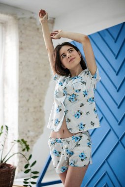 Genç, zayıf esmer bir kadın, beyaz pijama giyiyor, mavi çiçekli, sabahları esniyor. Beyaz tül ve mavi duvarlı büyük pencerenin yanındaki aydınlatma odasında. Uyku giysisi tasarımı.