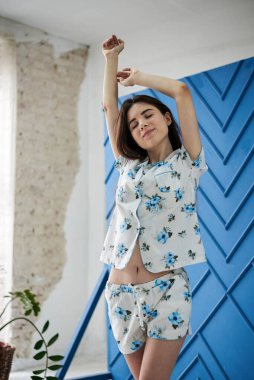 Genç, zayıf esmer bir kadın, beyaz pijama giyiyor, mavi çiçekli, sabahları esniyor. Beyaz tül ve mavi duvarlı büyük pencerenin yanındaki aydınlatma odasında. Uyku giysisi tasarımı.