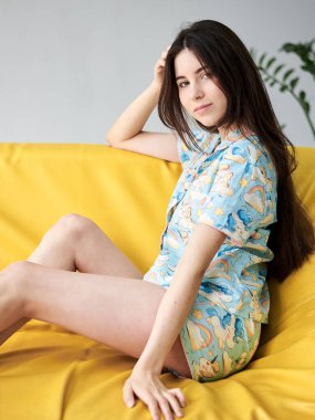 Renkli mavi pijama giyen, tek boynuzlu at desenli, sarı kanepede oturan, açık renk pencereli, zayıf, esmer bir kadın. Uyku giysisi tasarımı. Sabahları tatlı bir kız..