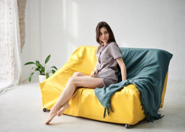 Gri saten pijama giyen, genç, zayıf, esmer bir kadın, yeşil kanepenin üzerinde beyaz tüllü büyük pencereli yeşil bir battaniye ile oturuyor. Uyku giysisi tasarımı. Sabahları tatlı bir kız..