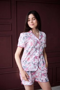 Genç, zayıf, esmer, renkli, açık pembe pijamalı, çiçek desenli gri pijamalı bir kadın kırmızı şarap duvarının önünde poz veriyor. Uyku giysisi tasarımı. Sabahları tatlı bir kız..