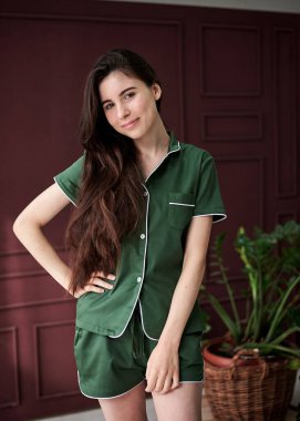 Koyu yeşil zümrüt pijama giyen, kırmızı bordo bordo duvarının önünde poz veren zayıf, esmer bir kadın. Uyku giysisi tasarımı. Sabahları tatlı bir kız. Noel sabahı. Pijama Partisi.