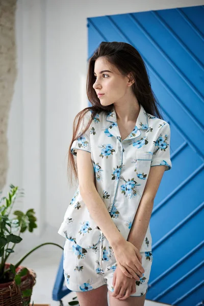 Açık mavi çiçek desenli beyaz pijama giyen, mavi duvarın önünde poz veren ve saksıdaki yeşil bitkiler için poz veren genç, zayıf, esmer bir kadın. Uyku giysisi tasarımı. Sabahları tatlı bir kız..