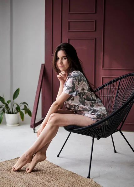 Genç, zayıf, esmer bir kadın. Renkli açık pembe ve çiçek desenli gri pijamalar giyiyor. Yeşil bitkilerle birlikte kırmızı duvarın yanındaki siyah koltukta oturuyor. Uyku giysisi tasarımı.