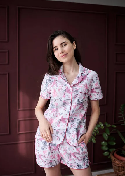 Genç, zayıf, esmer, renkli, açık pembe pijamalı, çiçek desenli gri pijamalı bir kadın kırmızı şarap duvarının önünde poz veriyor. Uyku giysisi tasarımı. Sabahları tatlı bir kız..