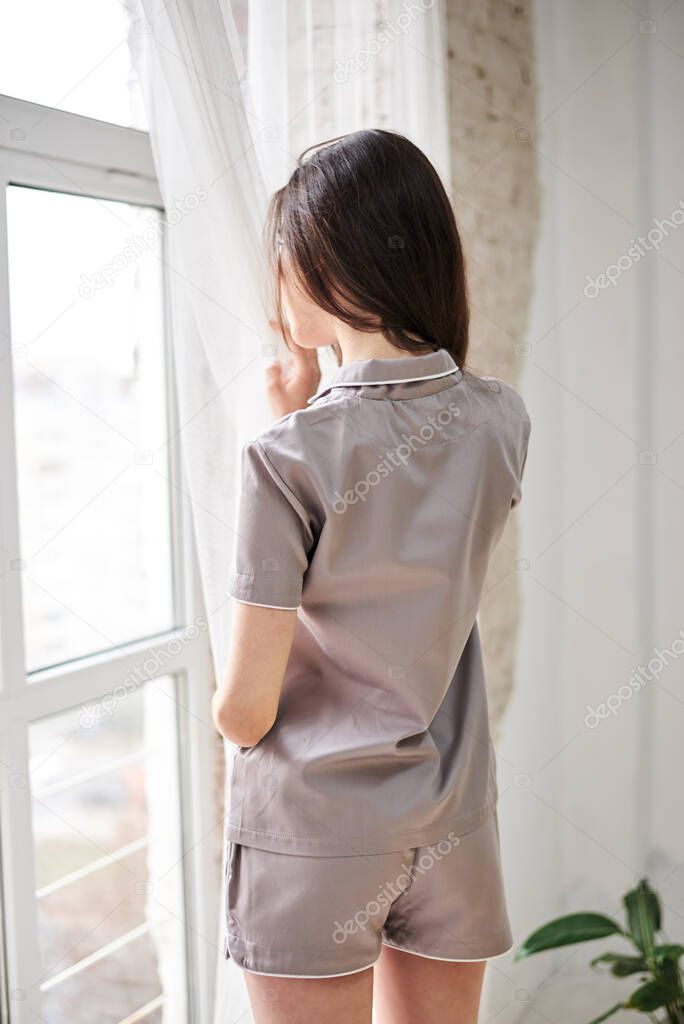 Young pretty slim brunette woman, wearing grey satin pajamas with, with her back to camera, looking through the window with white tulle, holding grey coffee cup mug, drinking. Sleepwear design.