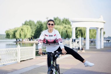 Sep 7, 2019-Ternopil / Ukrayna: Siyah tayt ve beyaz kazak giyen genç esmer kadın, yazın yeşil ağaçlı şehir parkında bisiklet sürüyor. Bisiklet süren bir kızın portresi. Sağlıklı yaşam konsepti