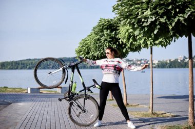 Sep 7, 2019-Ternopil / Ukrayna: Siyah tayt ve beyaz eşofman altı giyen, bisiklet tutan genç esmer kadın. Yaz sabahı göl kenarındaki şehir parkında bisiklet sürme sporu. Sağlıklı aktif yaşam konsepti.
