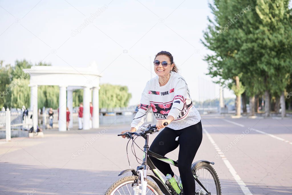 Sep 7, 2019-Ternopil/Ukraine:Young brunette woman, wearing black leggings and white pullover, riding a bike in city town park with green trees in summer.Portrait of girl, cycling. Healthy life concept