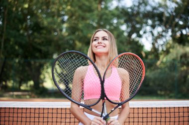 Genç sarışın kadın, pembe bluzlu beyaz etekli, kortta elinde turuncu zeminli iki tenis raketi tutuyor. Sporcunun dörtte üçü dışarıda antrenman yapıyor. Aktif sağlıklı yaşam.