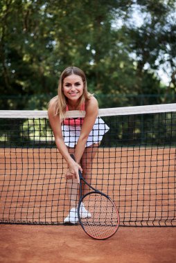 Genç sarışın kadın, pembe bluz, beyaz etek ve spor ayakkabı giyiyor tenis kortunda tenis ağına yaslanıyor, raket tutuyor, gülümsüyor... Tenis kıyafetli sporcu kadın portresi. Sağlıklı aktif yaşam tarzı kavramı.