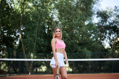 Genç sarışın kadın, pembe bluz ve beyaz etek giyiyor, tenis raketi tutuyor ve yazın kortta turuncu zeminli açık yeşil top tutuyor. Formda sporcu kadın portresi, dışarıda antrenman. Aktif sağlıklı yaşam