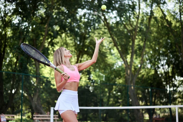ピンクのトップと白のスカートとスニーカーを身に着けている若いブロンドの女性は 夏にコートでテニスをします 4分の3のスポーツ女性の肖像画 テニスラケットとライトグリーンボールで外でトレーニング — ストック写真