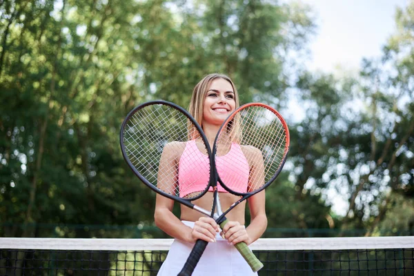 年轻的金发女子 身穿粉色上衣和白色裙子 夏天在球场上拿着两个有橙色场地的网球拍 四分之三的女运动员肖像 在外面训练 积极健康的生活 — 图库照片