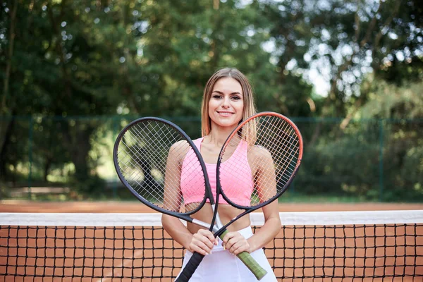 ピンクのトップスと白のスカートを着た若いブロンドの女性は 夏にオレンジの地面とコートで2つのテニスラケットを保持しています スポーツ女性の4分の3の肖像画 外でのトレーニング 健康的な生活 — ストック写真