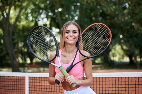 ピンクのトップスと白のスカートを着た若いブロンドの女性は 夏にオレンジの地面とコートで2つのテニスラケットを保持しています スポーツ女性の4分の3の肖像画 外でのトレーニング 健康的な生活 — ストック写真