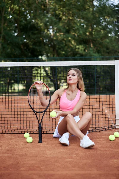 若いブロンドの女性は ピンクの上 白いスカートとスニーカーを着て テニスネットにもたれて座って 訓練後にリラックスした オレンジ色の地面とテニスコートのスポーツウーマンの肖像 健康な生活 — ストック写真