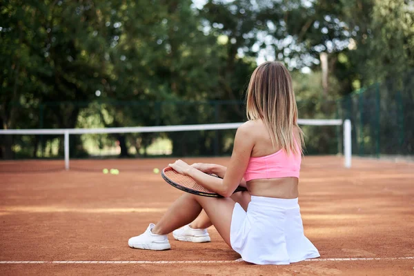 若いブロンドの女性 ピンクの上を身に着けています 白いスカートとスニーカー テニスコートに座って ラケットを保持 トレーニング後にリラックス オレンジ色の地面とテニスコートのスポーツウーマンの肖像 健康な生活 — ストック写真