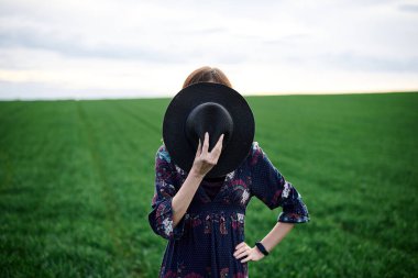 Genç sarışın kadın, uzun koyu renk boho elbisesi giyiyor, baharda yeşil tarlada siyah şapka takıyor. Dışarıda çayırda poz veren bir model. Doğal ortamda hippi müzisyen..