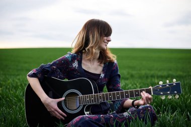 Genç sarışın kadın, uzun koyu boho elbisesi giyiyor, baharda yeşil çayırlarda oturuyor. Dışarıda çayırda poz veren bir model. Doğal ortamda hippi müzisyen..