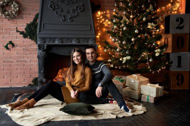 Gri kazaklı yakışıklı esmer adam ve hardal sarısı örülü elbise tutan, sarılan genç kadın. Aşık bir çift şöminenin yanında halıda oturuyor ve karanlık odada tuğladan duvarlı Noel ağacı..