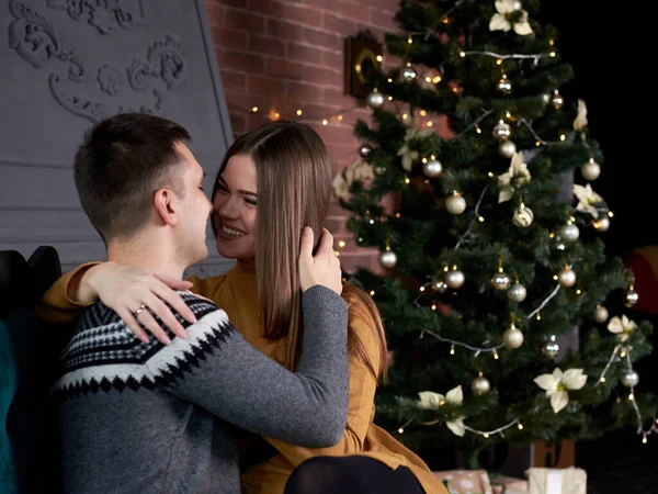 Young couple in love sitting by fireplace near Christmas tree, kissing on a date. Handsome brunette man, wearing grey winter sweater kissing pretty woman in mustard yellow knitted dress.