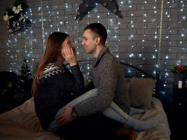 愛の若いカップルは 背後にクリスマスライトとベッドの上に座って 新しい年の前夜に暗い部屋に抱きつく灰色の冬のセーターを着て ブルネットの男性と女性 ロマンチックなクリスマス — ストック写真