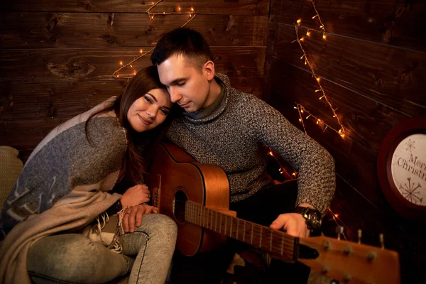 クリスマスの新しい年の木製の部屋小屋のロマンチックなデートを愛する若いカップル前夜 ブルネット男 グレーの冬のセーターを着て 暖かい毛布で覆われた美しい女性のための演奏ギターを保持 — ストック写真