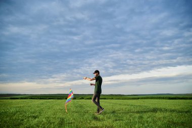 Genç, esmer, sıska, koyu yeşil tişörtlü, yeşil çayır çimenliğinde renkli uçurtmayla oynayan adam. Mavi bulutlu gökyüzünde uçan uçurtma. Aile eğlencesi aktivitesi