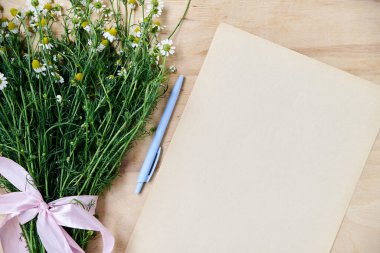 Pembe kurdeleli küçük papatya buketi ve açık mavi kalemli bej kağıt çarşaf ahşap masada. Vahşi çiçeklerle dolu doğal bir arka plan. Şiirler, mektuplar, romantik notlar için ekolojik doku.