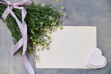 Küçük papatya buketi pembe kurdeleyle bağlanmış ve gri ahşap masa üzerinde bej kağıt çarşaf var. Vahşi çiçeklerin doğal arka plan resmi. Şiirler, mektuplar, romantik notlar için ekolojik doku.