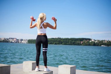 Siyah tayt ve beyaz bluz giyen sarışın genç bir kadın beton platformda şehir dışında antrenman yapıyor ve kırmızı ağırlıklar taşıyor. Yazın kadın güç eğitimi. Sağlıklı yaşam tarzı kavramı.