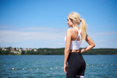 Siyah tayt ve beyaz bluz giyen genç sarışın kadın, arka planda mavi güneşli gökyüzü ile şehir gölü kenarında poz veriyor. Yazın kadın fitness eğitimi. Sağlıklı yaşam tarzı kavramı.