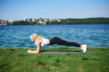 Siyah tayt ve beyaz bluz giyen genç sarışın kadın yaz sabahı gölün kenarında gri yoga minderinde tahta egzersizleri yapıyor. Kadın sporu eğitimi dışarıda, temiz havada. Sağlıklı yaşam tarzı.