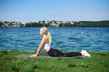 Siyah tayt ve beyaz bluz giyen genç sarışın kadın, yaz sabahı göl kenarında gri yoga minderinde esneme egzersizleri yapıyor. Kadın sporu eğitimi dışarıda, temiz havada. Sağlıklı yaşam tarzı.