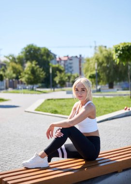 Sarışın, beyaz bluzlu ve siyah taytlı genç bir kadın güneşli bir yaz sabahında şehir parkında bankta oturuyor. Dışarıdaki fitness eğitiminden sonra dinlenen bir kadın. Sağlıklı yaşam tarzı kavramı.
