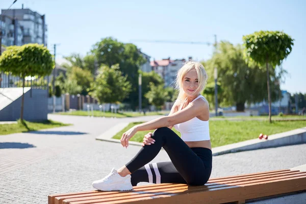 夏日阳光灿烂的早晨 身穿白色上衣和黑色腿的金发美女坐在城市公园的长椅上 女性在户外运动后休息 健康生活方式概念 — 图库照片