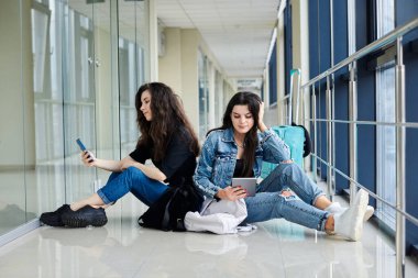 İki genç esmer kız, açık hava koridorunda arkada bavullarıyla oturuyorlar, telefon ve tableti kontrol ediyorlar, günlük kot giyiyorlar. Kız arkadaşlar, hava yoluyla seyahat ediyorlar, uçmayı bekliyorlar..