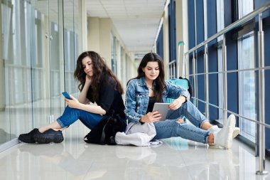 İki genç esmer kız, açık hava koridorunda arkada bavullarıyla oturuyorlar, telefon ve tableti kontrol ediyorlar, günlük kot giyiyorlar. Kız arkadaşlar, hava yoluyla seyahat ediyorlar, uçmayı bekliyorlar..