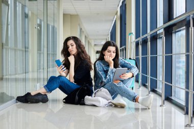 İki genç esmer kız, açık hava koridorunda arkada bavullarıyla oturuyorlar, telefon ve tableti kontrol ediyorlar, günlük kot giyiyorlar. Kız arkadaşlar, hava yoluyla seyahat ediyorlar, uçmayı bekliyorlar..