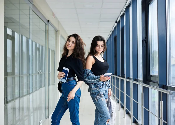 两名年轻的黑发女孩站在有巨大窗户的轻型机场走廊里 穿着休闲装 持有国际护照和登机证 女朋友坐飞机旅行 — 图库照片
