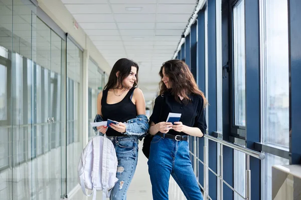 2人の若いブルネットの女の子は 巨大な窓のある軽い空港の廊下に立って カジュアルなジーンズの服を着て 国際パスポートを保持し 搭乗券を渡します 飛行機で旅する彼女たち — ストック写真