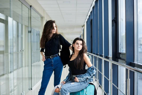 Two young brunette girls, sitting on small mint carry on luggage in light airport hallway with huge windows, wearing casual jeans clothes. Girlfriends, traveling by air, waiting for flight.