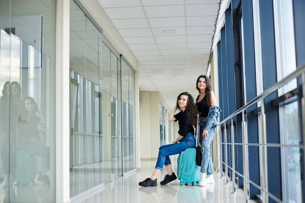 Two young brunette girls, sitting on small mint carry on luggage in light airport hallway with huge windows, wearing casual jeans clothes. Girlfriends, traveling by air, waiting for flight.