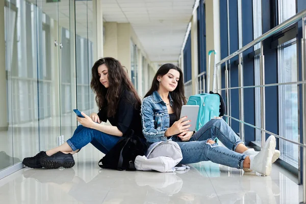 İki genç esmer kız, açık hava koridorunda arkada bavullarıyla oturuyorlar, telefon ve tableti kontrol ediyorlar, günlük kot giyiyorlar. Kız arkadaşlar, hava yoluyla seyahat ediyorlar, uçmayı bekliyorlar..